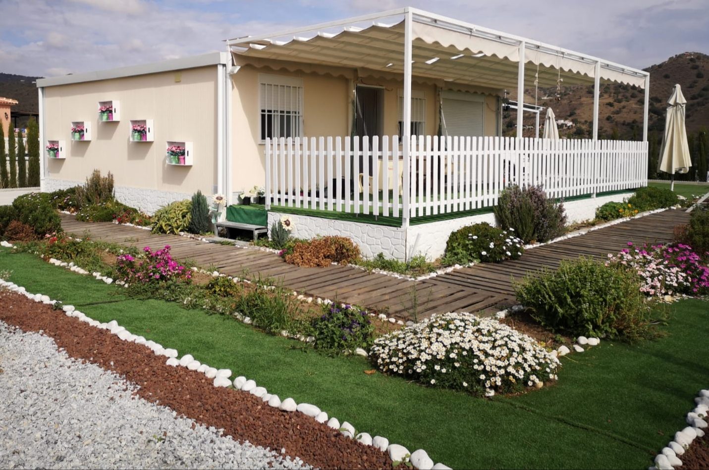 Casa modular color crema con pérgola, porche vallado y jardín con flores